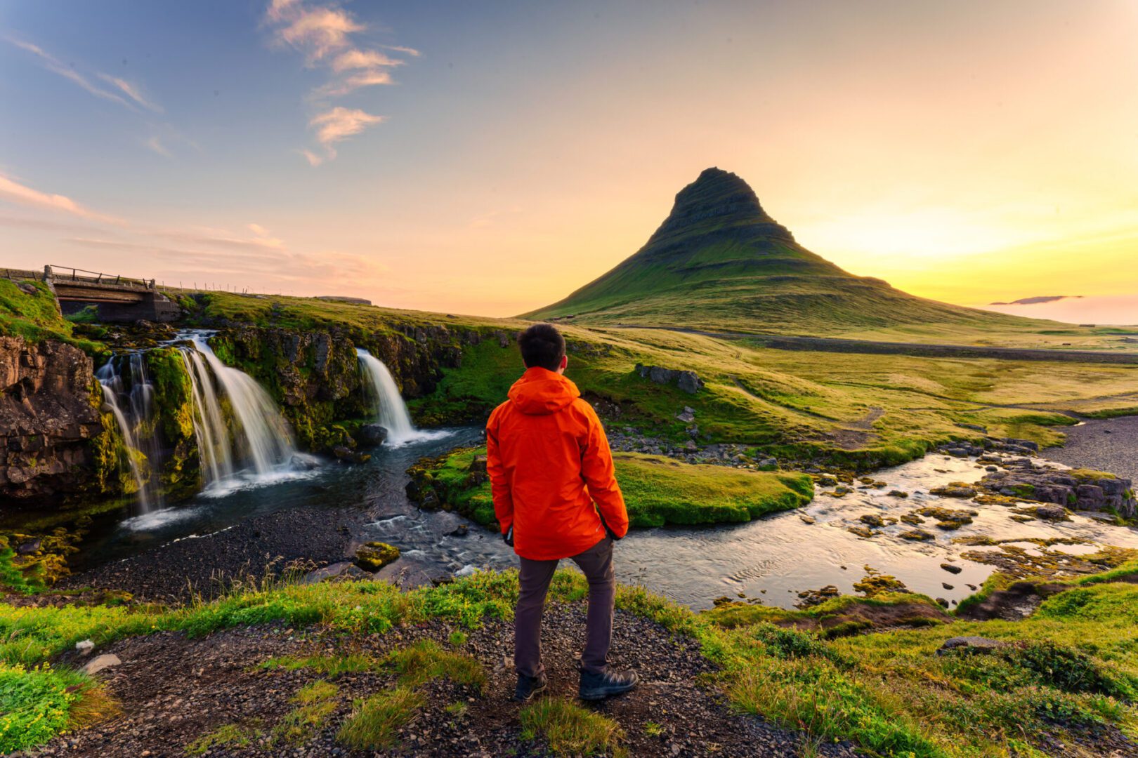 Rear of traveler man in red jacket standing with sunrise over Kirkjufell mountain with Kirkjufellsfoss waterfall in summer at Snaefellsnes peninsula, Iceland